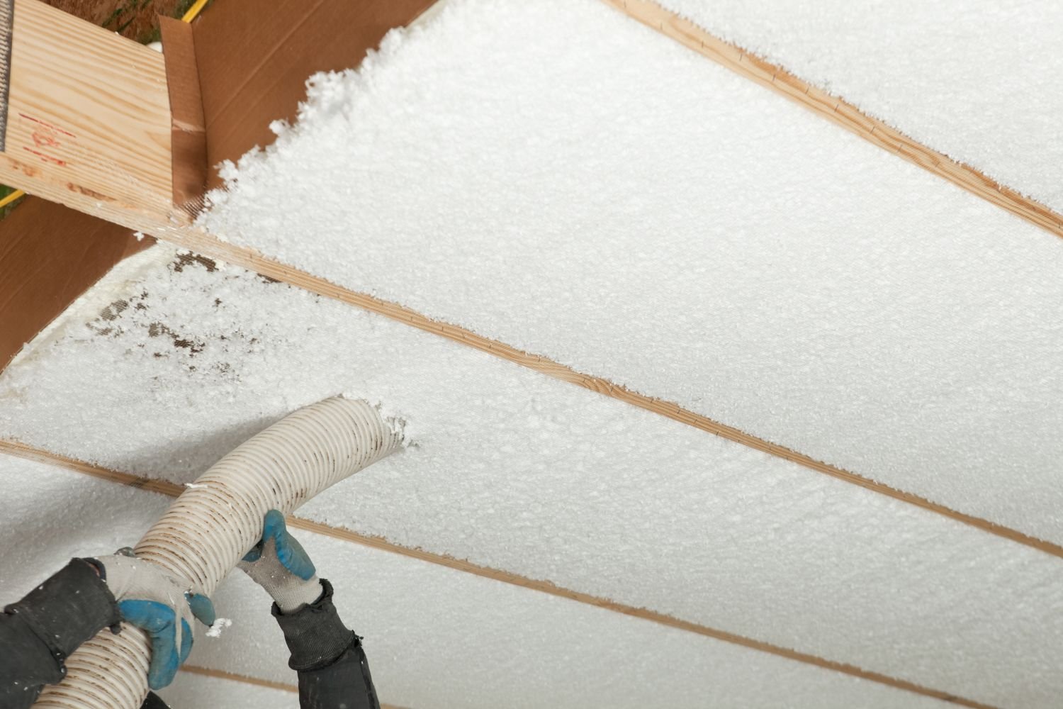 A worker uses a hose to blow insulation into a ceiling cavity, with wooden beams visible in the background.