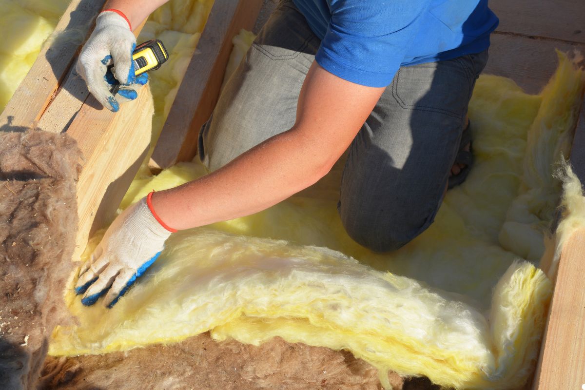 A man installs insulation in a house, carefully placing material between the wooden beams of the ceiling.