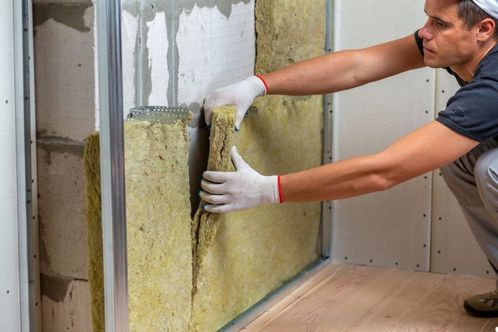 A man in a white shirt and gloves carefully installs insulation, ensuring a snug fit for energy efficiency in a construction area.