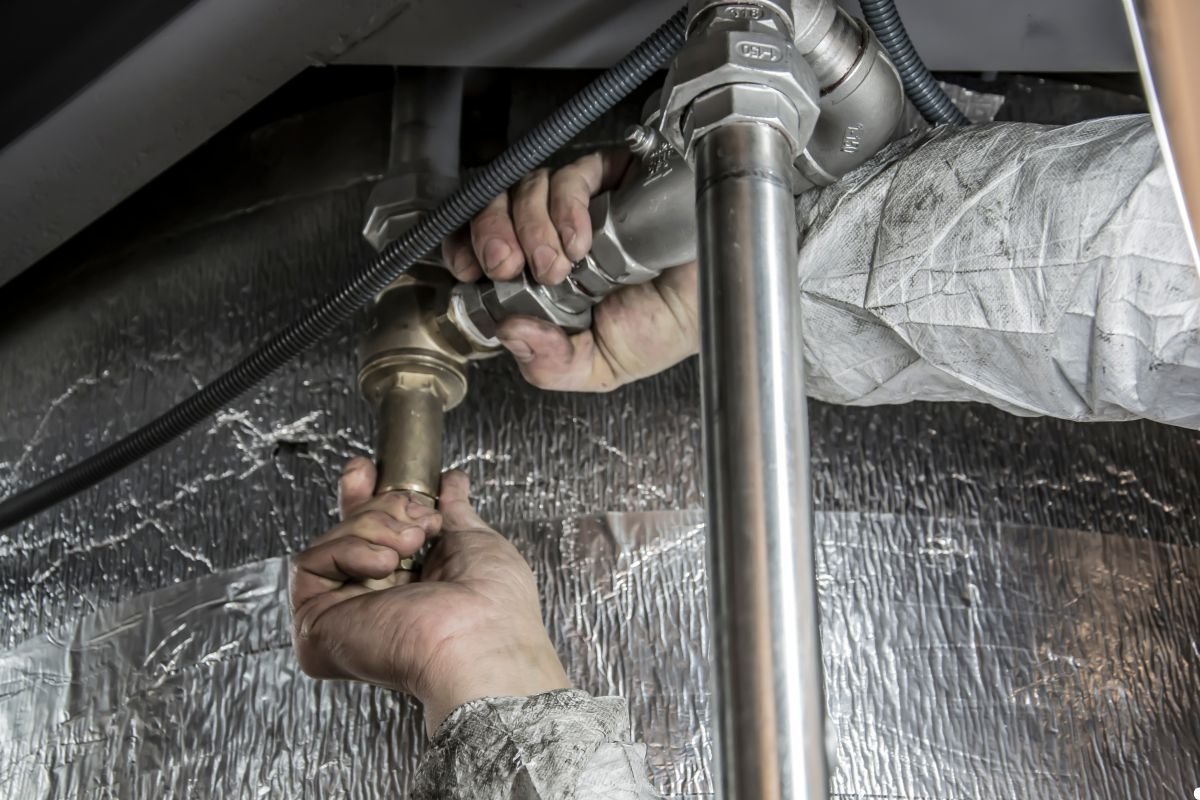 A man in a white coat repairs a pipe, focused on his task in a well-lit environment.