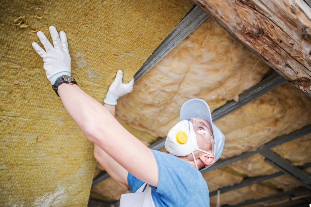 A man in a mask and gloves installs insulation, focusing on safety while working on a construction project.